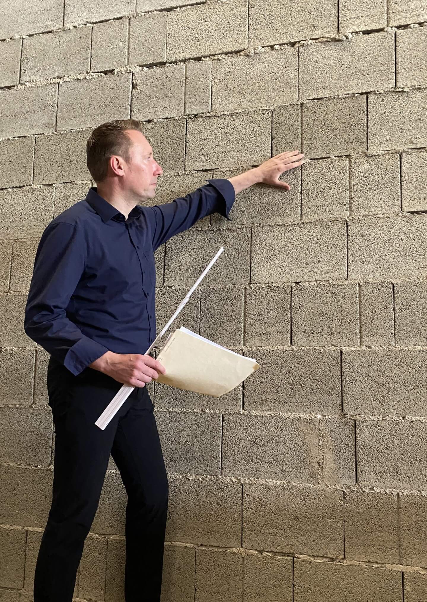 Baumeister DI Franz Grassl, Chef des Unternehmens, inspiziert eine Betonblockwand auf einer Baustelle. Er trägt ein dunkelblaues Hemd, hält Dokumente und zeigt mit der Hand auf die Wand.